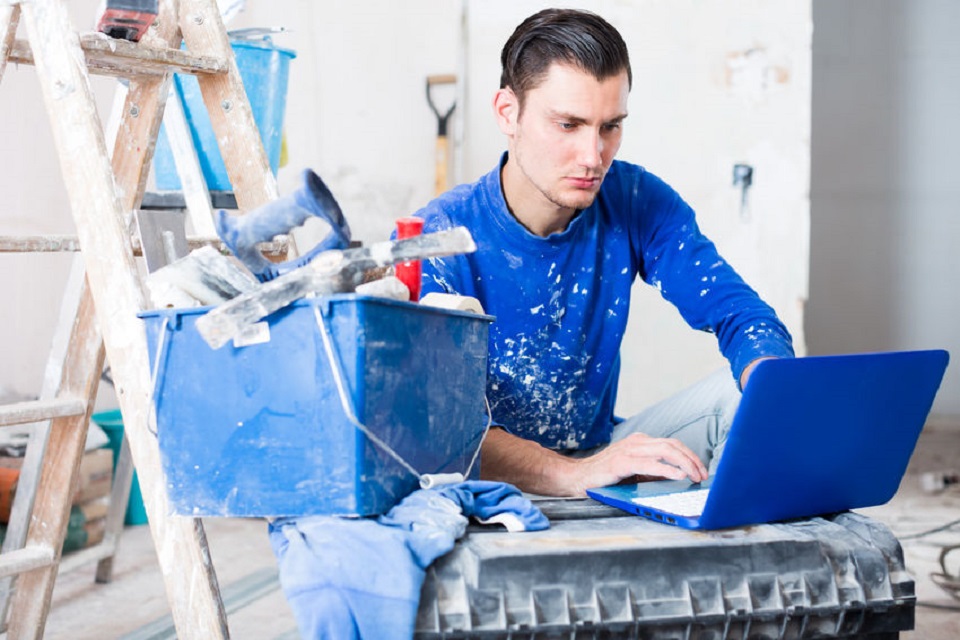 Young male contractor renovating house and working on laptop in repairable apartment